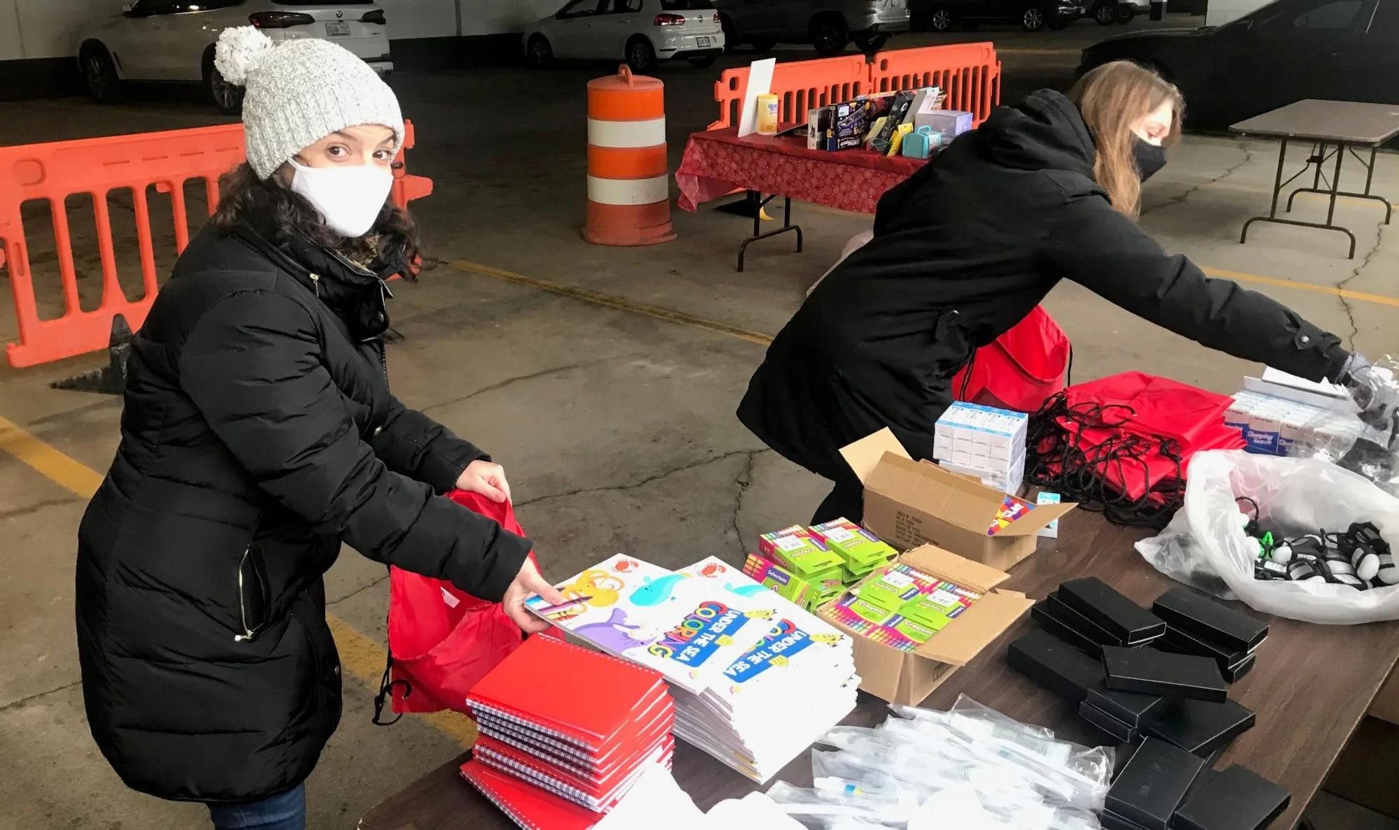 Two masked volunteers organizing backpacks, notebooks, educational materials, and supplies on tables in an indoor distribution area.