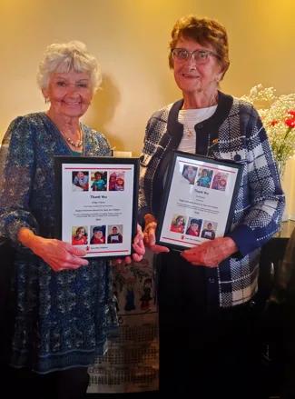 Two people standing indoors holding framed recognition certificates featuring photos of children, with floral arrangements and interior d?�cor visible in the background.