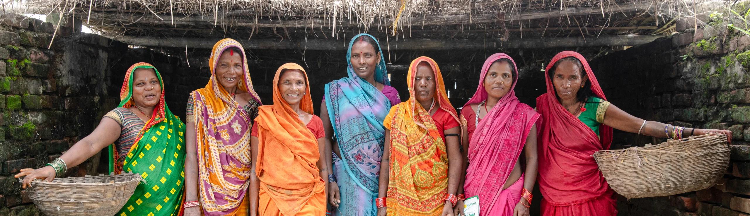 Group of women wearing colorful traditional clothing standing together in front of a thatched structure, with woven baskets visible on either side.