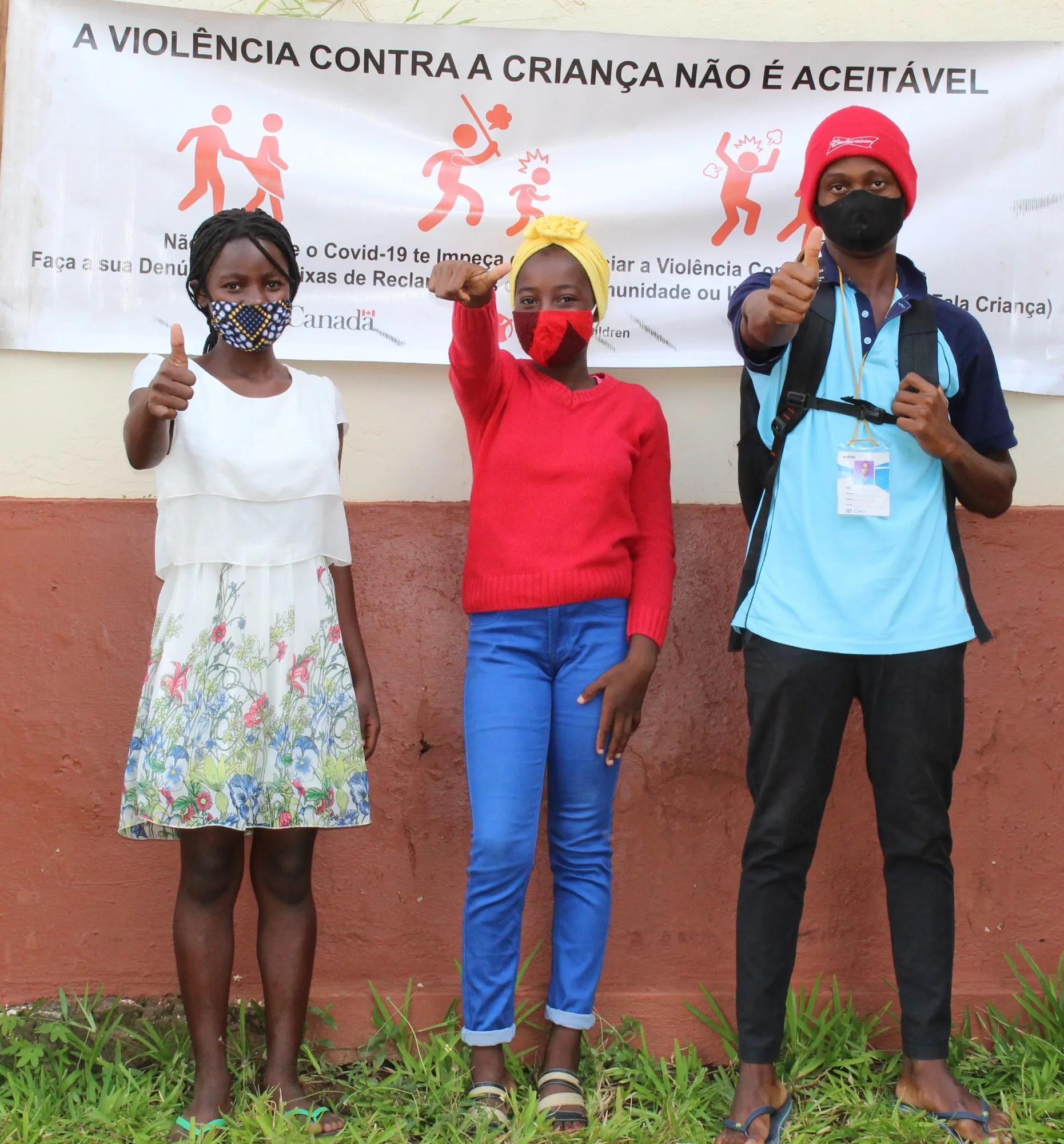 Three masked youths standing in front of a child protection awareness banner, giving thumbs-up gestures and pointing toward the camera outdoors.