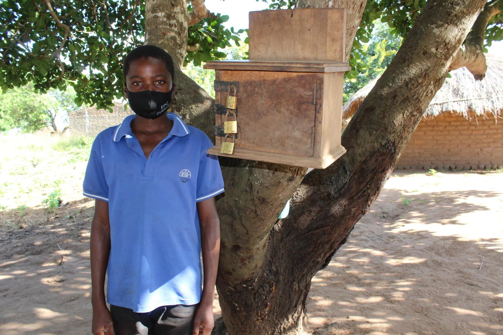 Young person wearing a face mask standing next to a locked wooden donation or savings box attached to a tree in a rural outdoor setting.