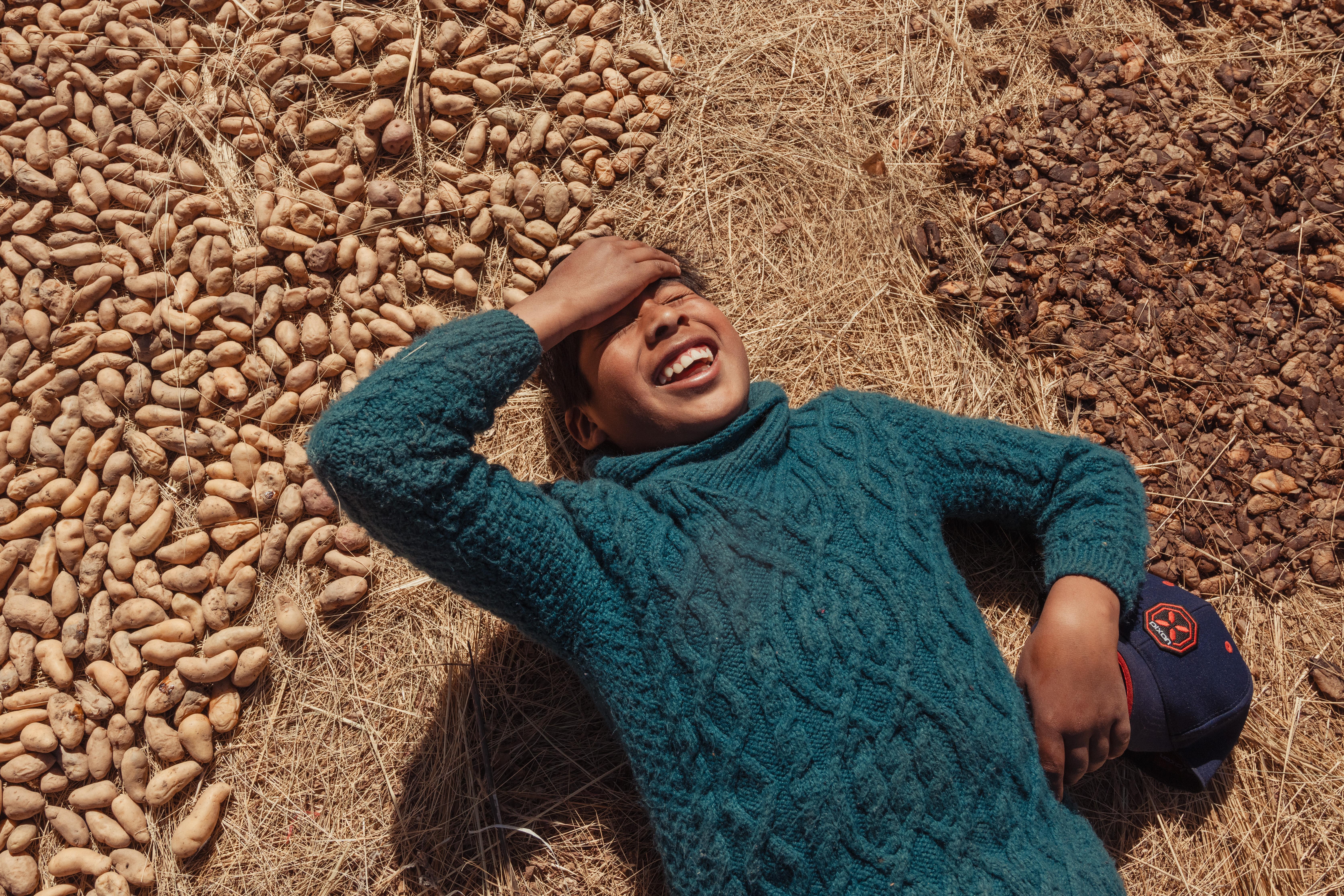 Jhon, 11, lies laughing on a pile of his family's potatoes drying in the sun, wearing a turquoise wool jumper