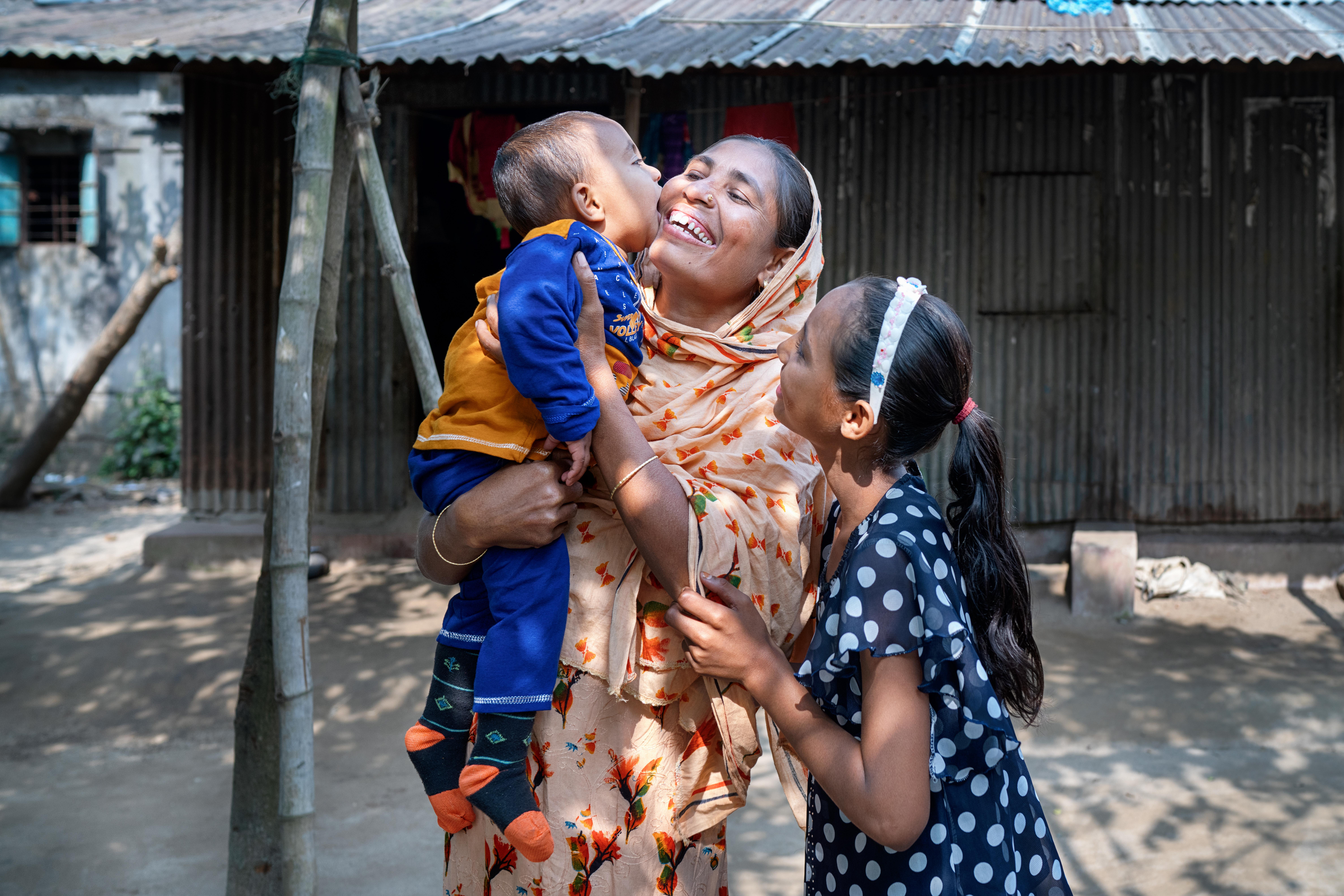 Rohima stands smiling outside her home in Sylhet, Bangladesh, with her children Nafiz, 14 months and Taiyeba, 10