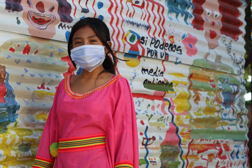 A young girl wears a face mask and looks to the camera, as she stand in front of a colourful wall of cartoon drawings 