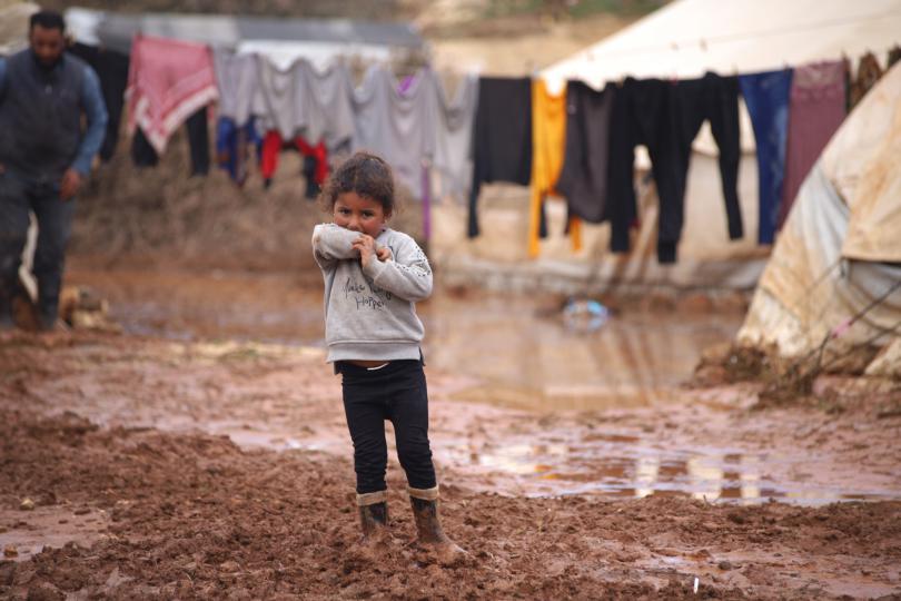 A girl standing in front of her family tent, through flood water and mud in a  camp for internally displaced people in Idlib, Syria