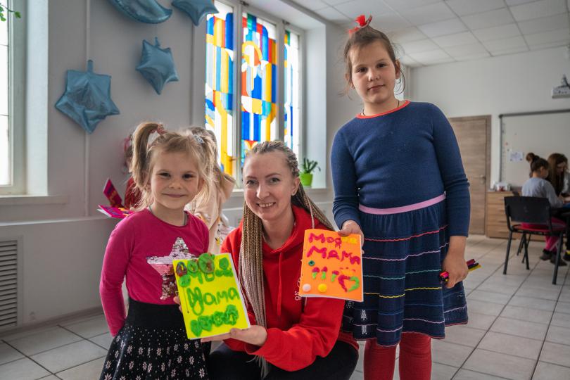 Olga Shults, Save the Children's programme manager, poses for portrait with *Marta (left) and *Lera (right) holding gift cards at Child Friendly Space in Mykolaiv, Ukraine.