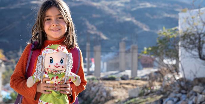 Aslı 9, with her favourite doll, stands in front of ruins after the February 2023 earthquakes in Türkiye