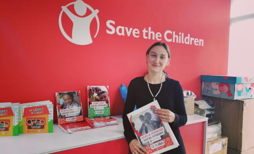 Ayse stands in front of the Save the Children logo, holding some of the books used in the temporary learning centre